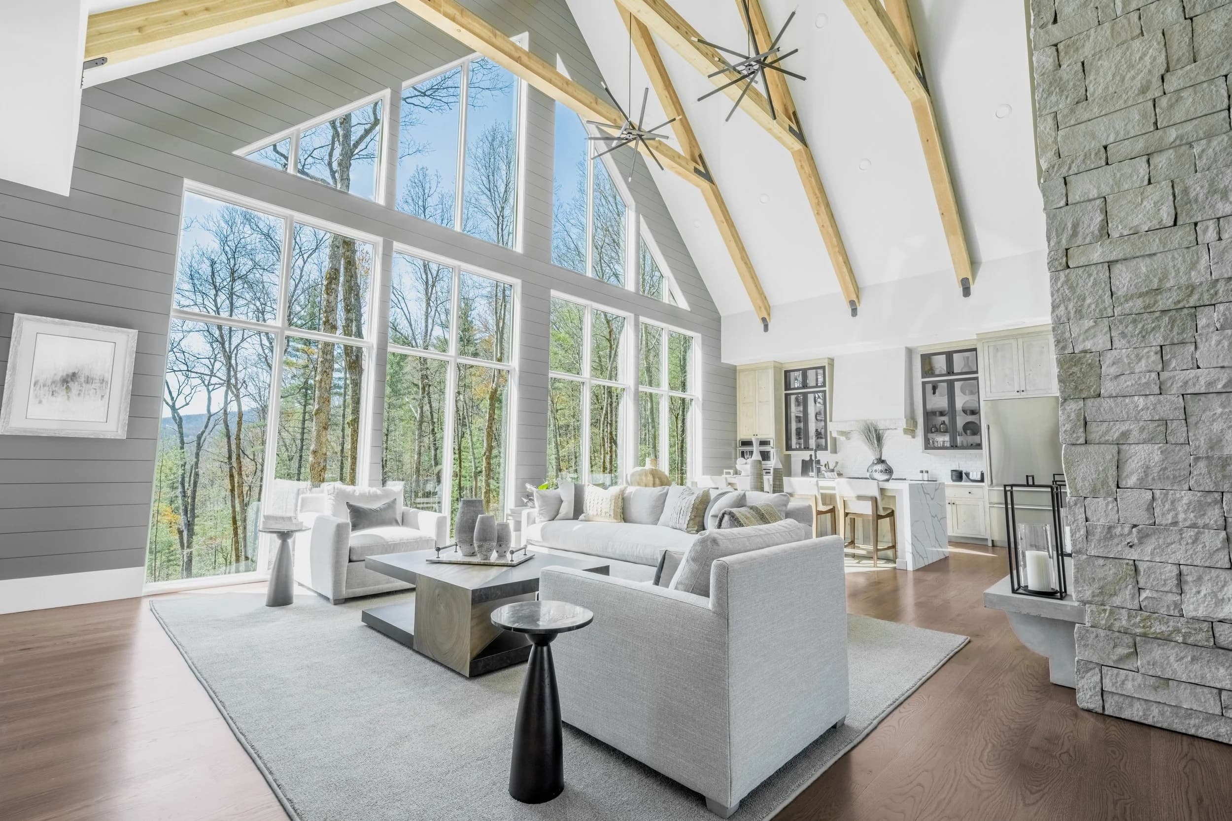 Great room with timber trusses, a stone fireplace, and a wall of windows looking into the trees.