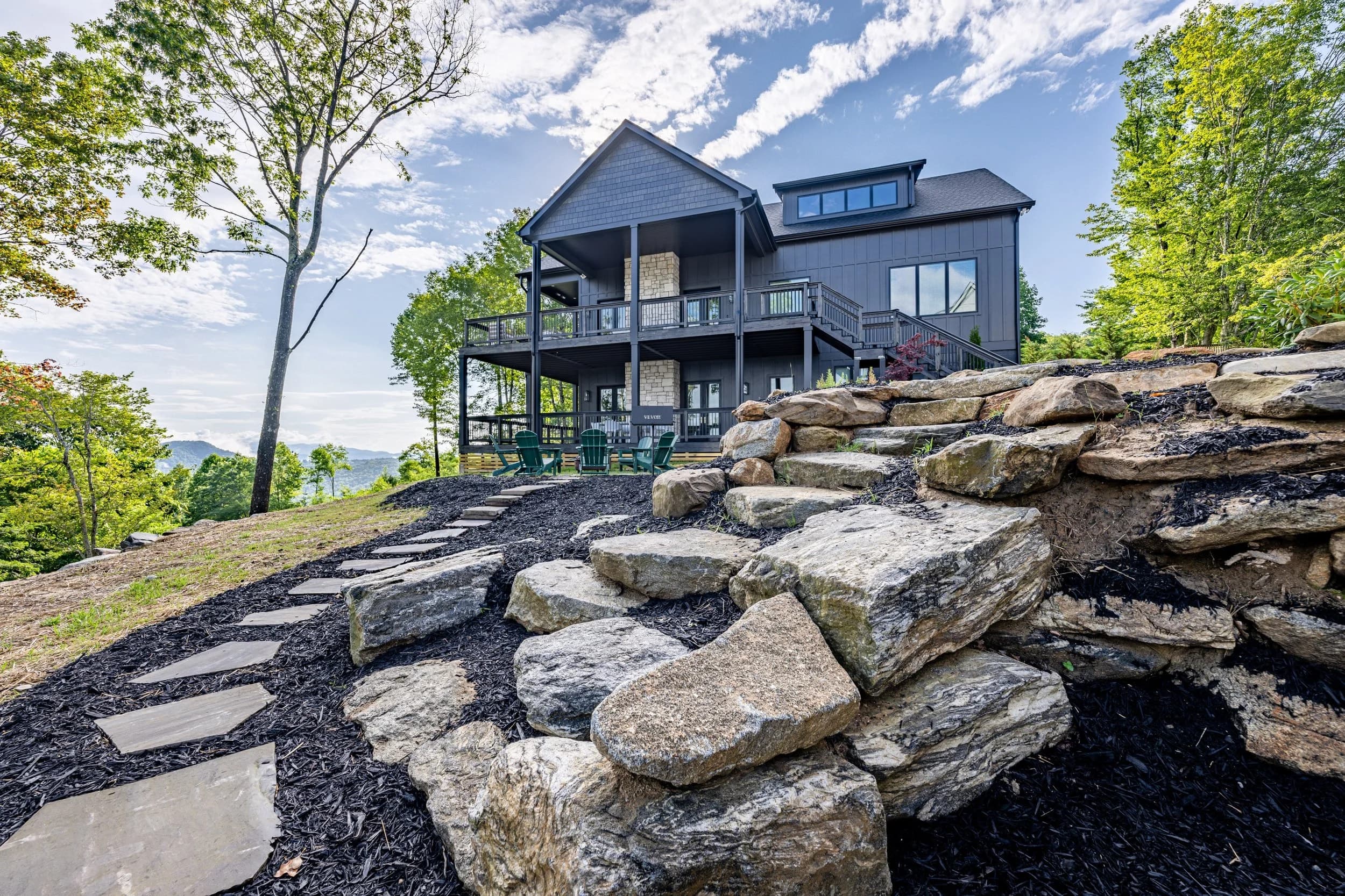 A custom home set into a wooded slope, with stone steps and a view toward the mountains.